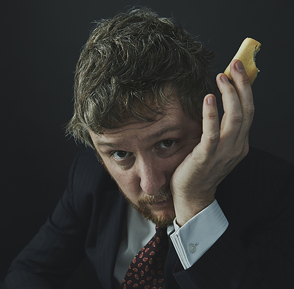 Portrait photograph of Tim Key gazing into the camera. He wears a suit and he holds a mug and biscuit as he leans over a newspaper.
