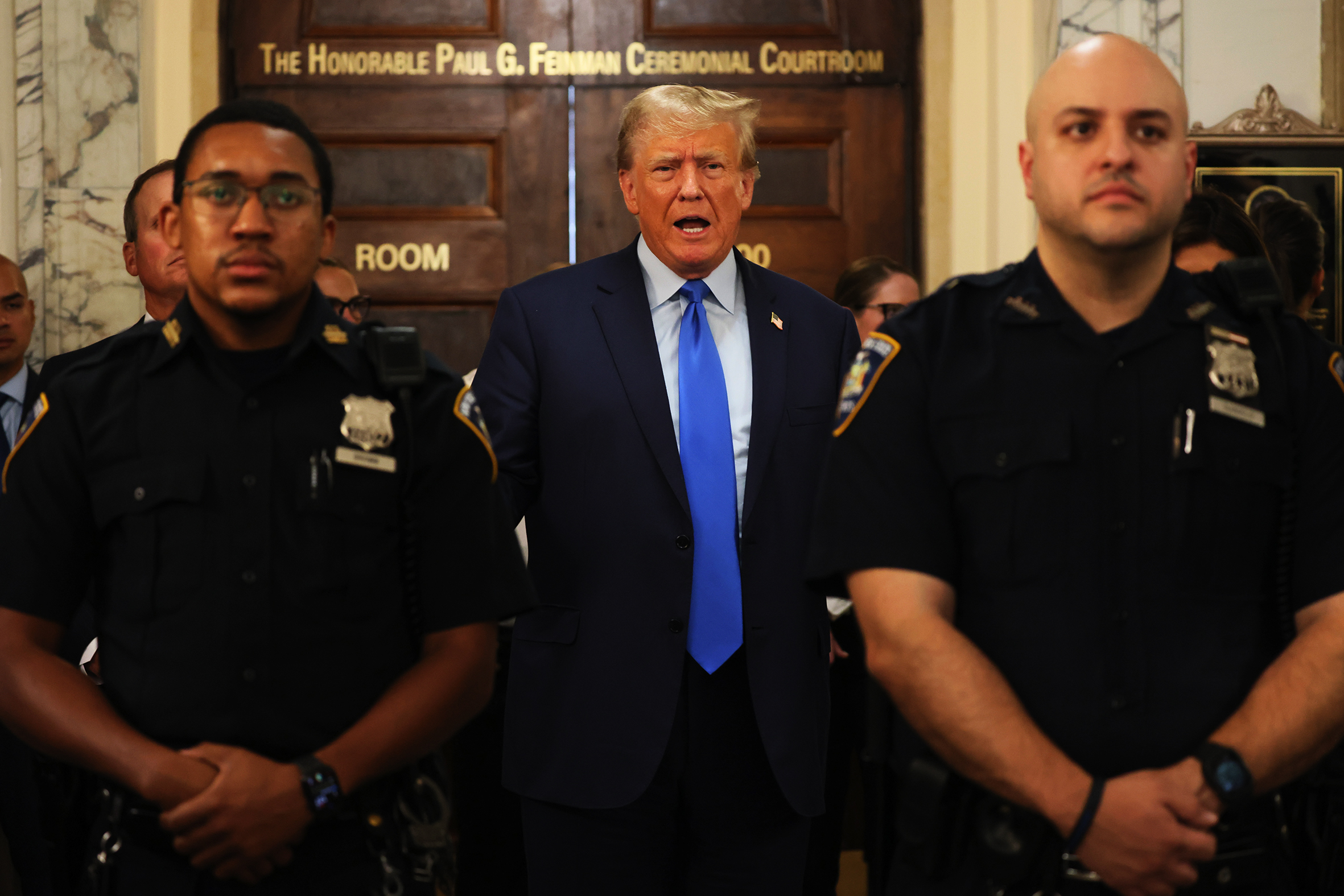 Donald Trump in a courthouse and flanked by two police officers. 