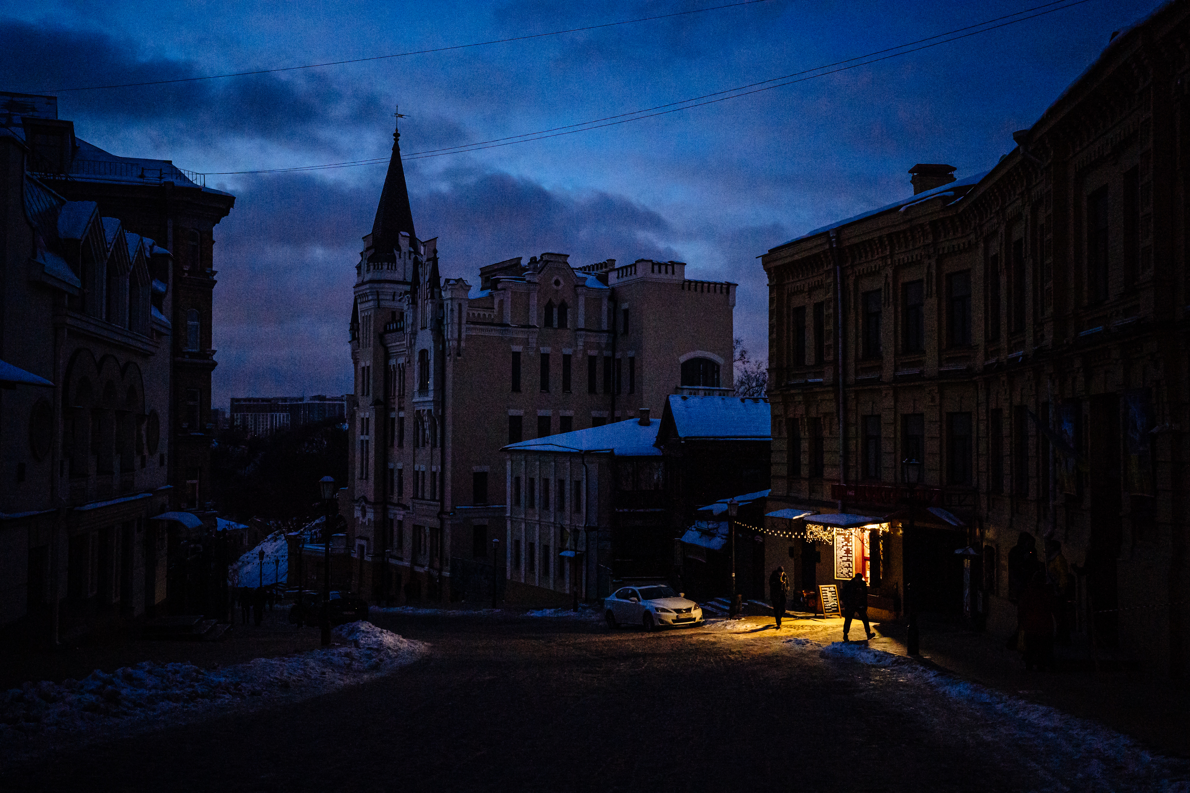 A dark street in what appears to be a European town with old buildings and snow scattered around on the rooftops; a single shopfront can be seen with yellow light spilling out onto the street.