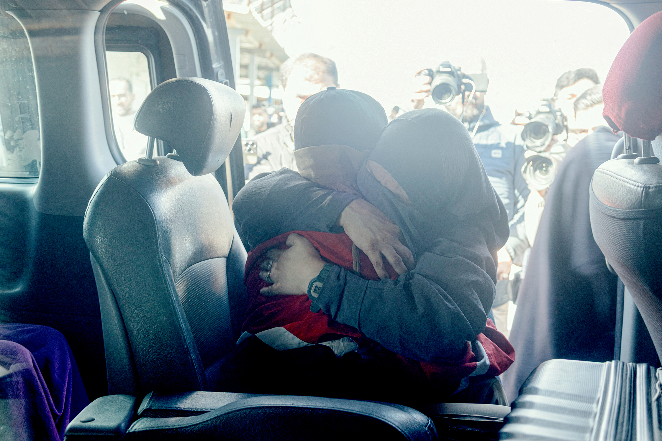 Two women embrace in a car with photographers behind them.