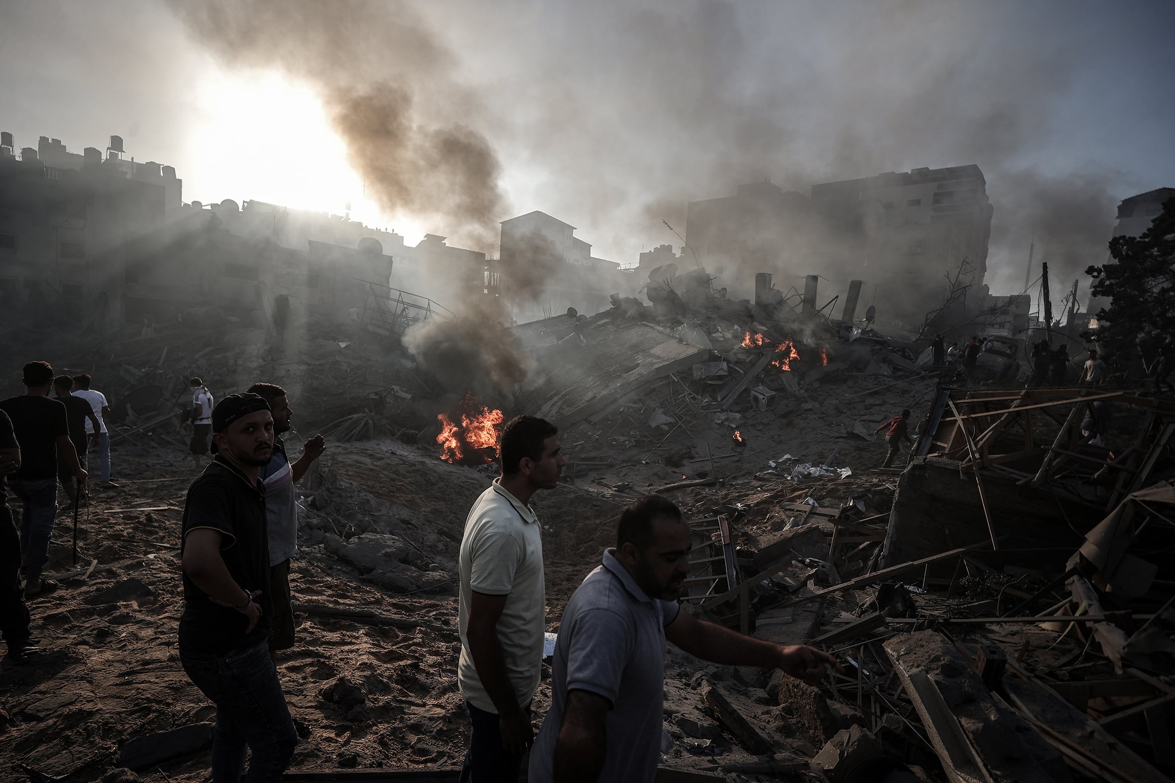 A group of men stand beside mountain of debris caused by air strikes. Smoke fills the sky.