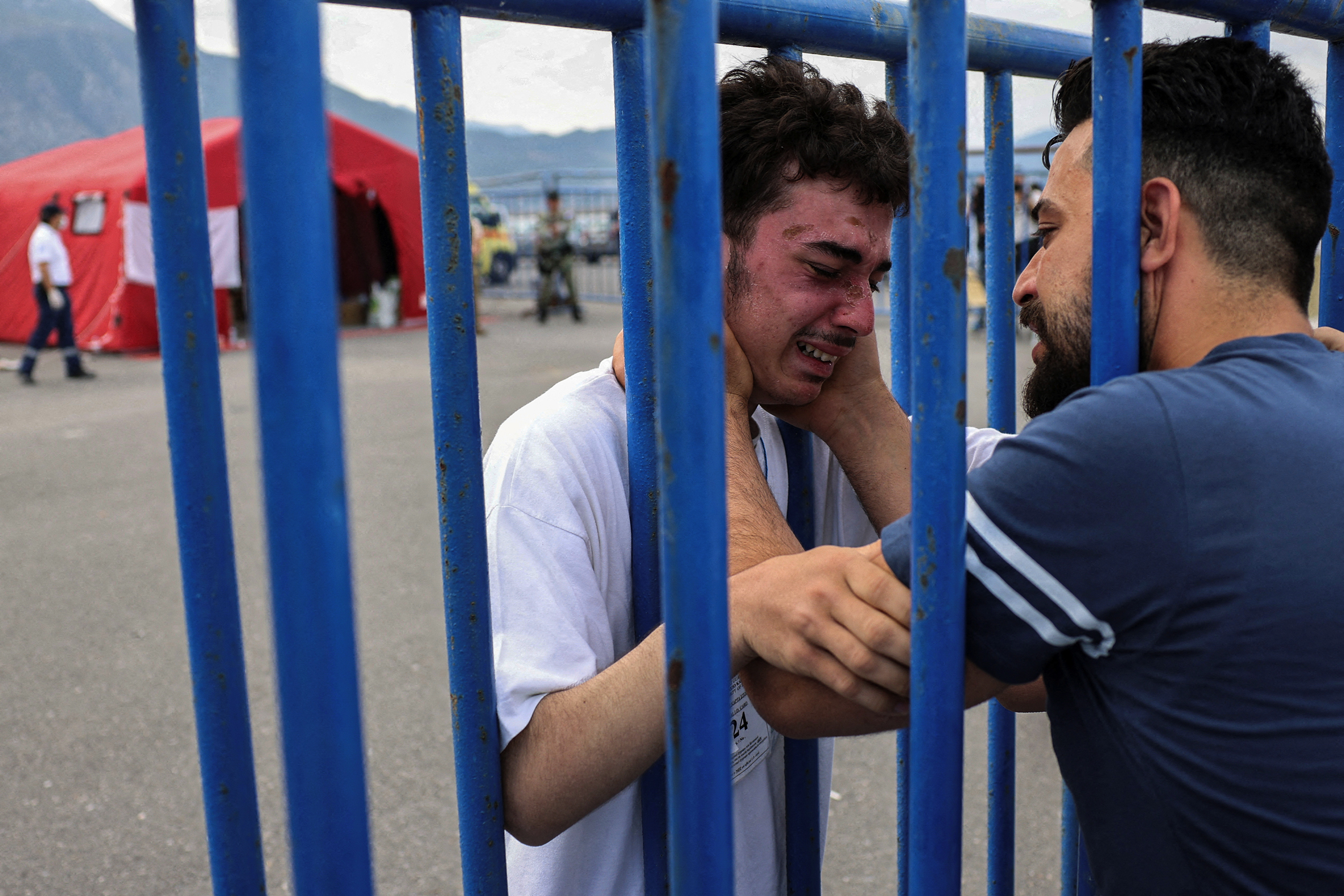 A man and teenage boy hold each other through steel bars. The teenage boy is distressed and has a severely sunburnt face.