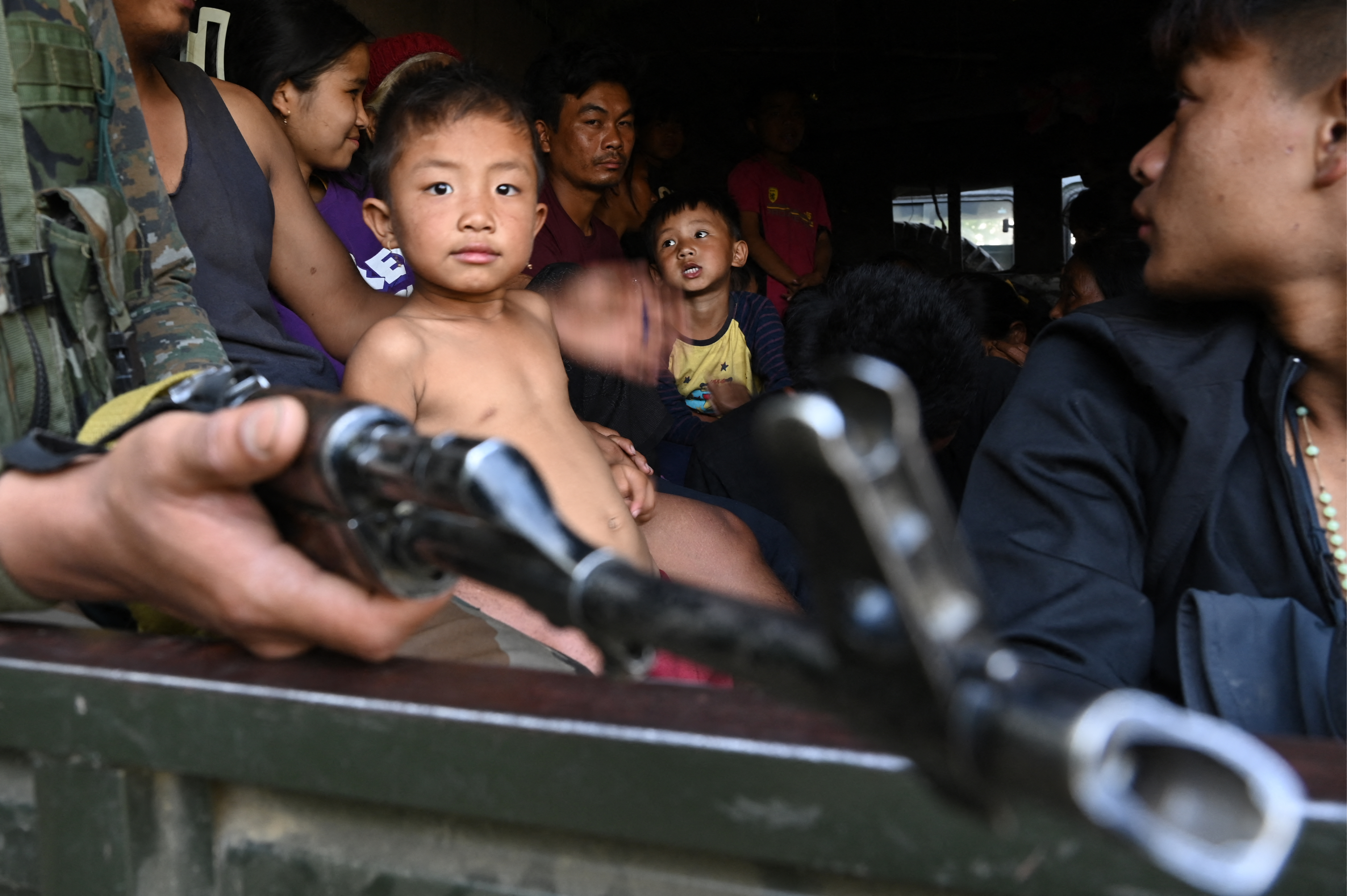 A family sits in a truck, a man's machine gun hanging out the window.
