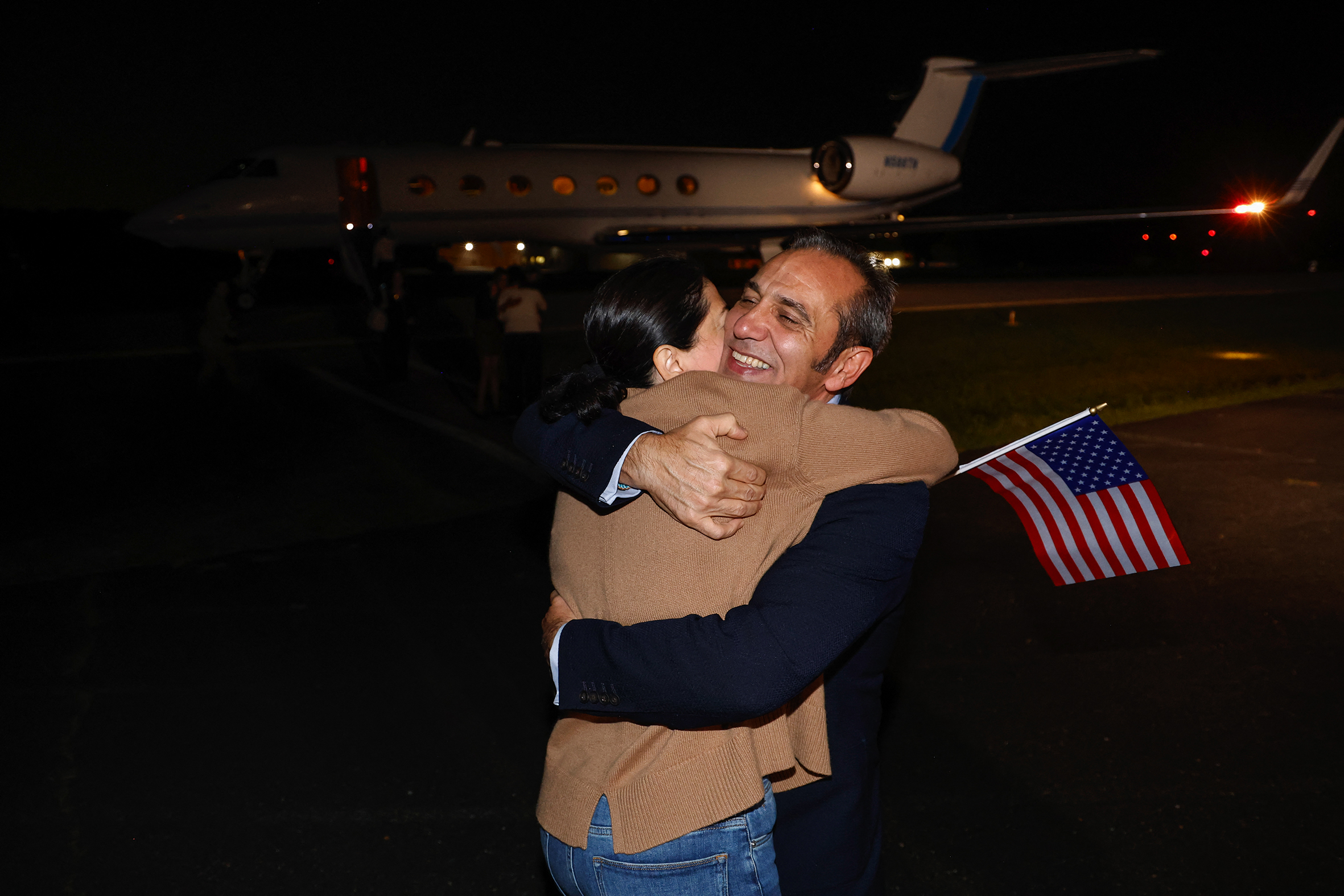 A man in a suit embraces a woman holding an American flag, with a jet in the background.