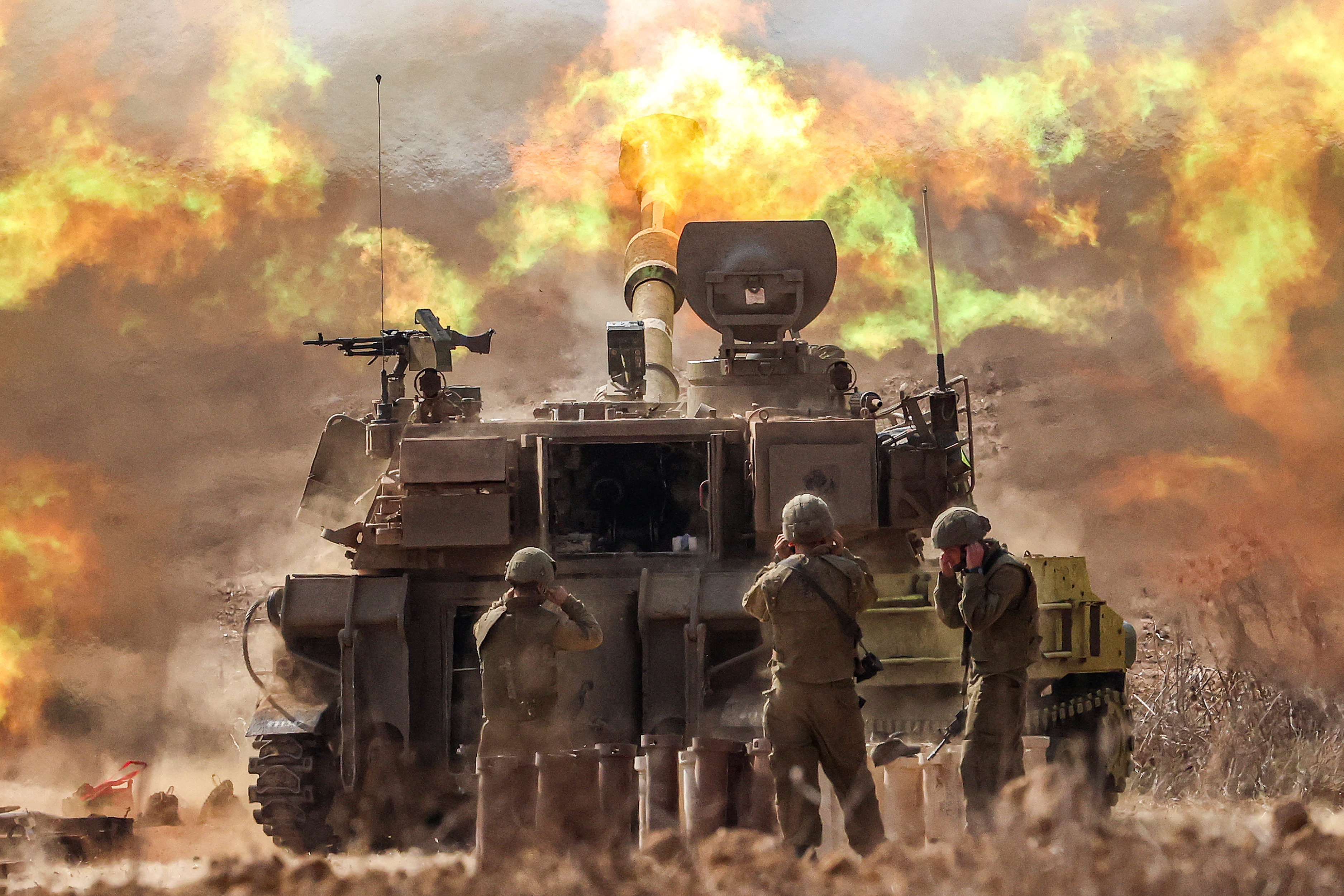 Soldiers stand behind a tank as it fires. Flames fill the sky.