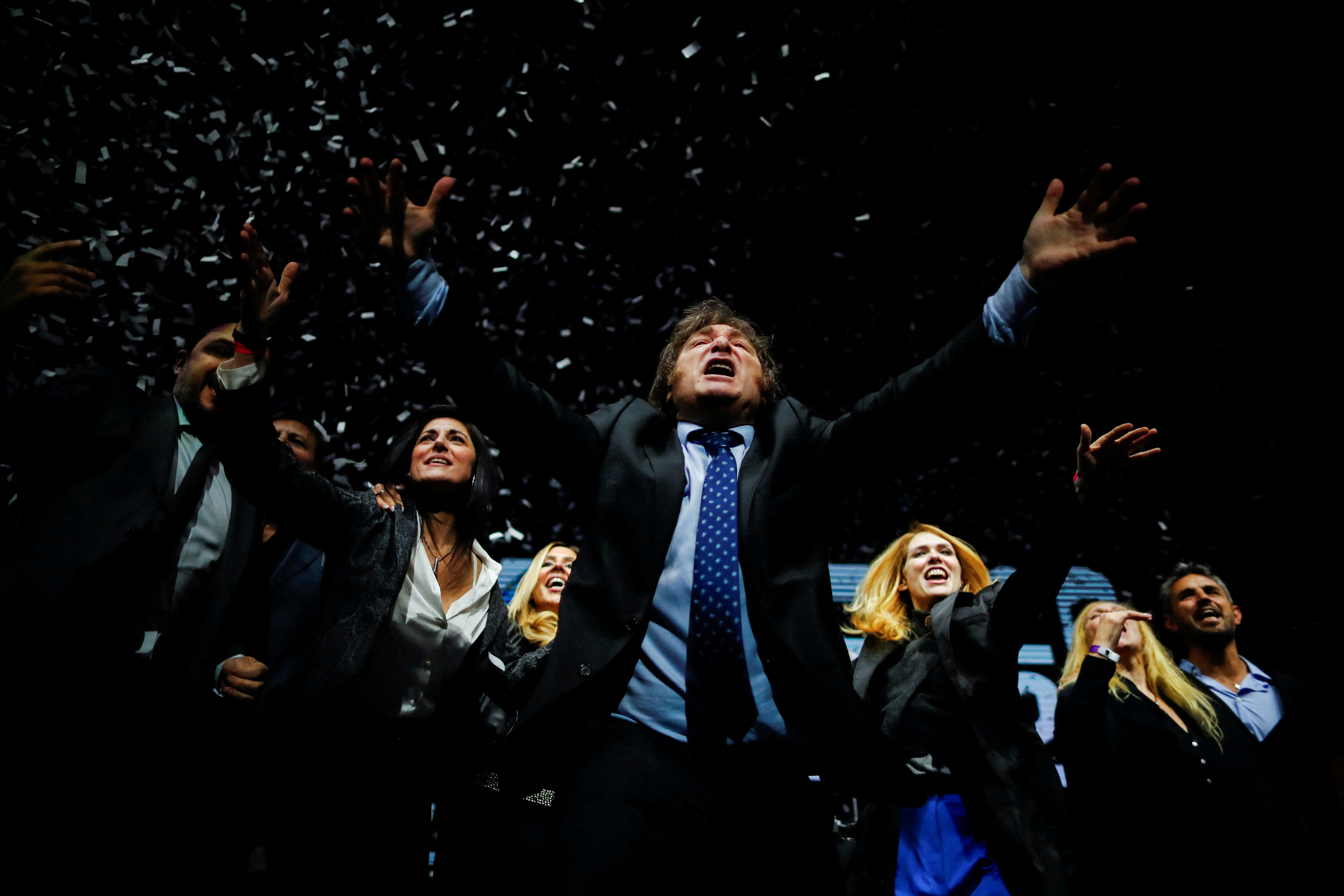 A middle-aged man with sideburns and dressed in a suit celebrates with confetti falling around him.