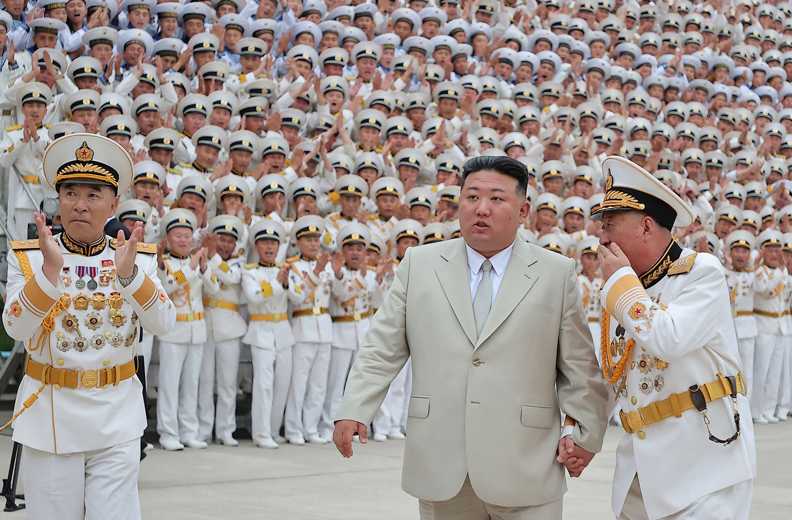 Kim Jong-un in a beige suitm holding hands with a navy officer in a white uniform. Behind them, a crowd in white uniform applaud.