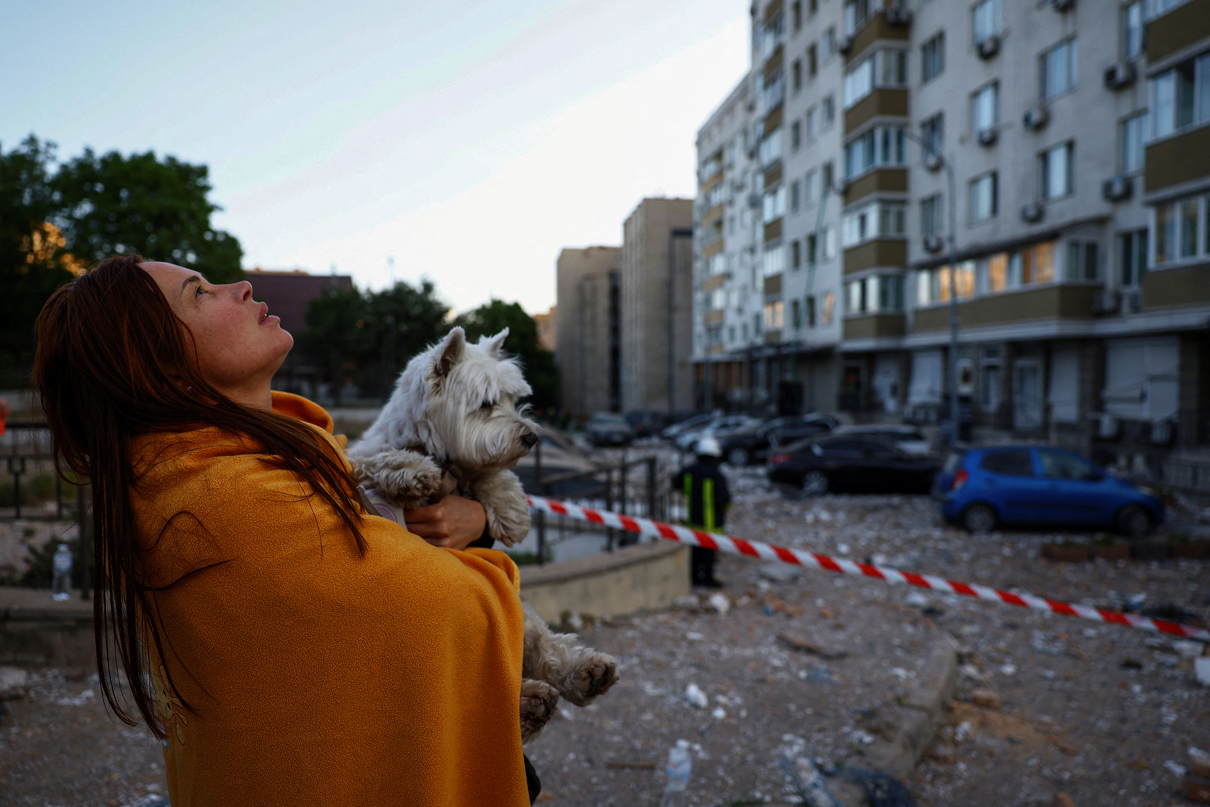 A woman stands with a blanket wrapped around her and a dog in her arms. City apartments surround her, with debris scattered across the street.