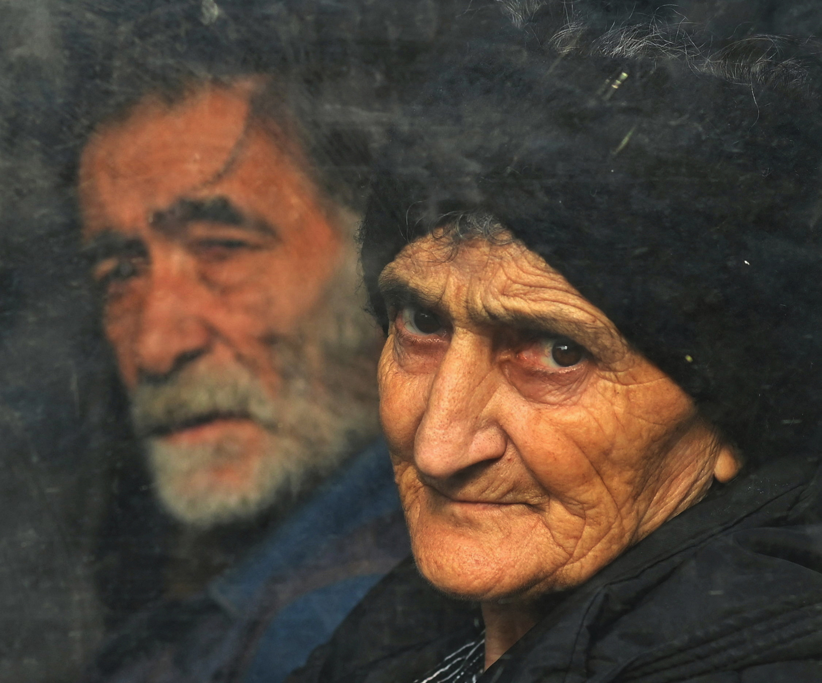 An elderly woman with an intense gaze looks through a dirty window.