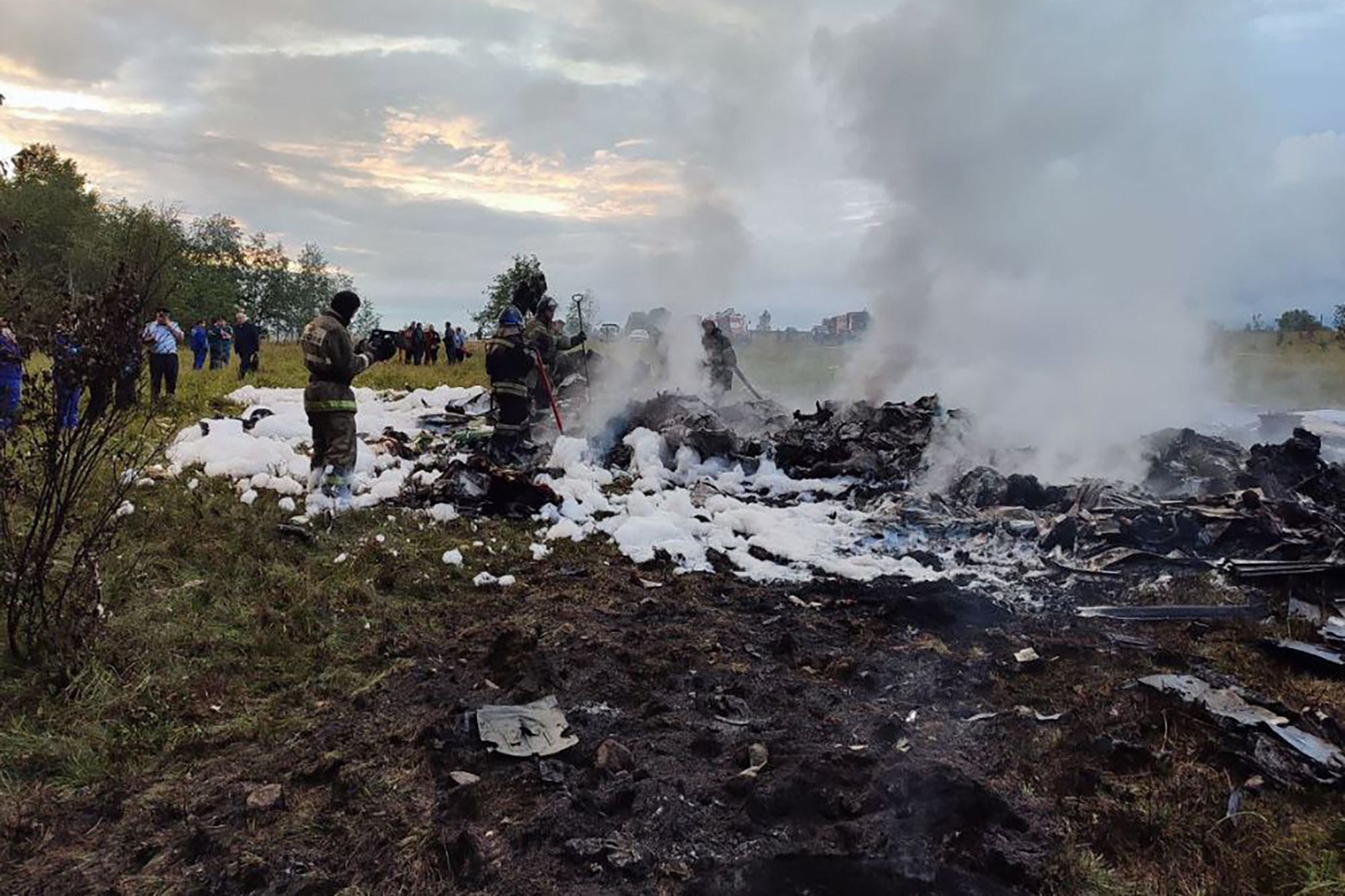 The site of a plane crash. Smoke rises from the ground as people stand around the debris with shovels.
