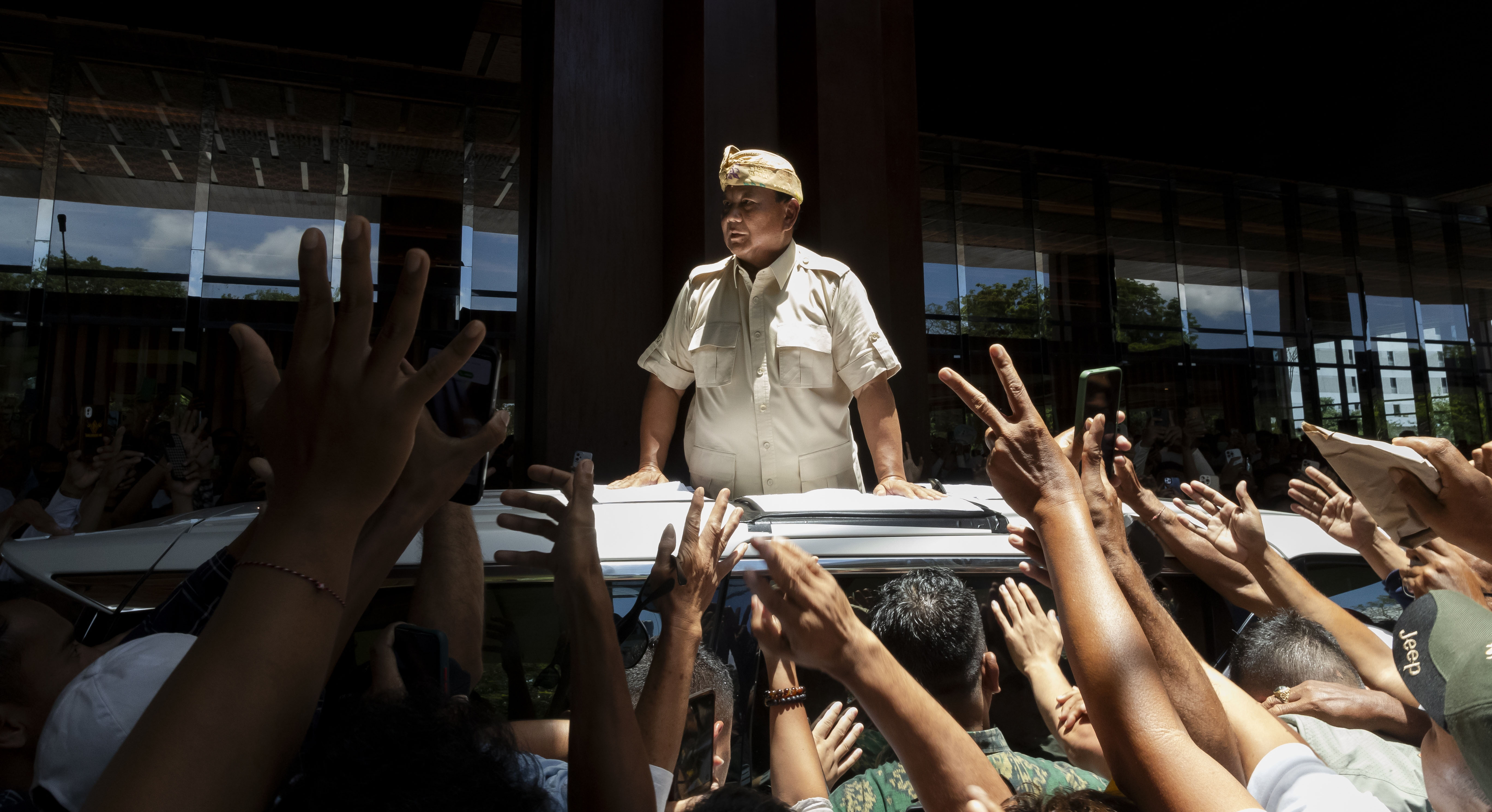 A man in stands through the sunroof of a car, overlooking a crowd that's reaching out to him.