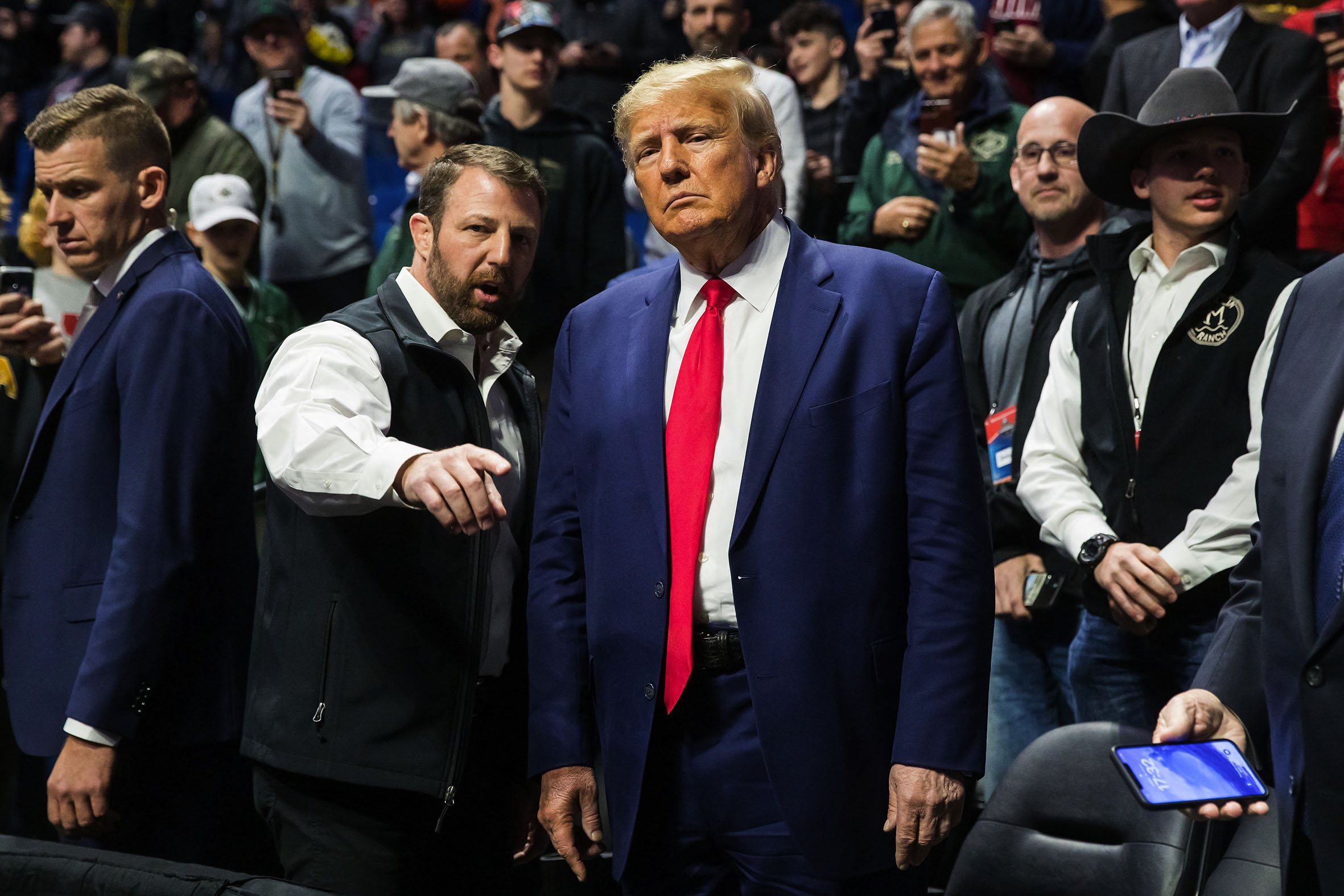 Former US president Donald Trump stands in a crowd and is wearing a navy blue suit with a red tie. The person beside him is pointing at something at something out of frame.