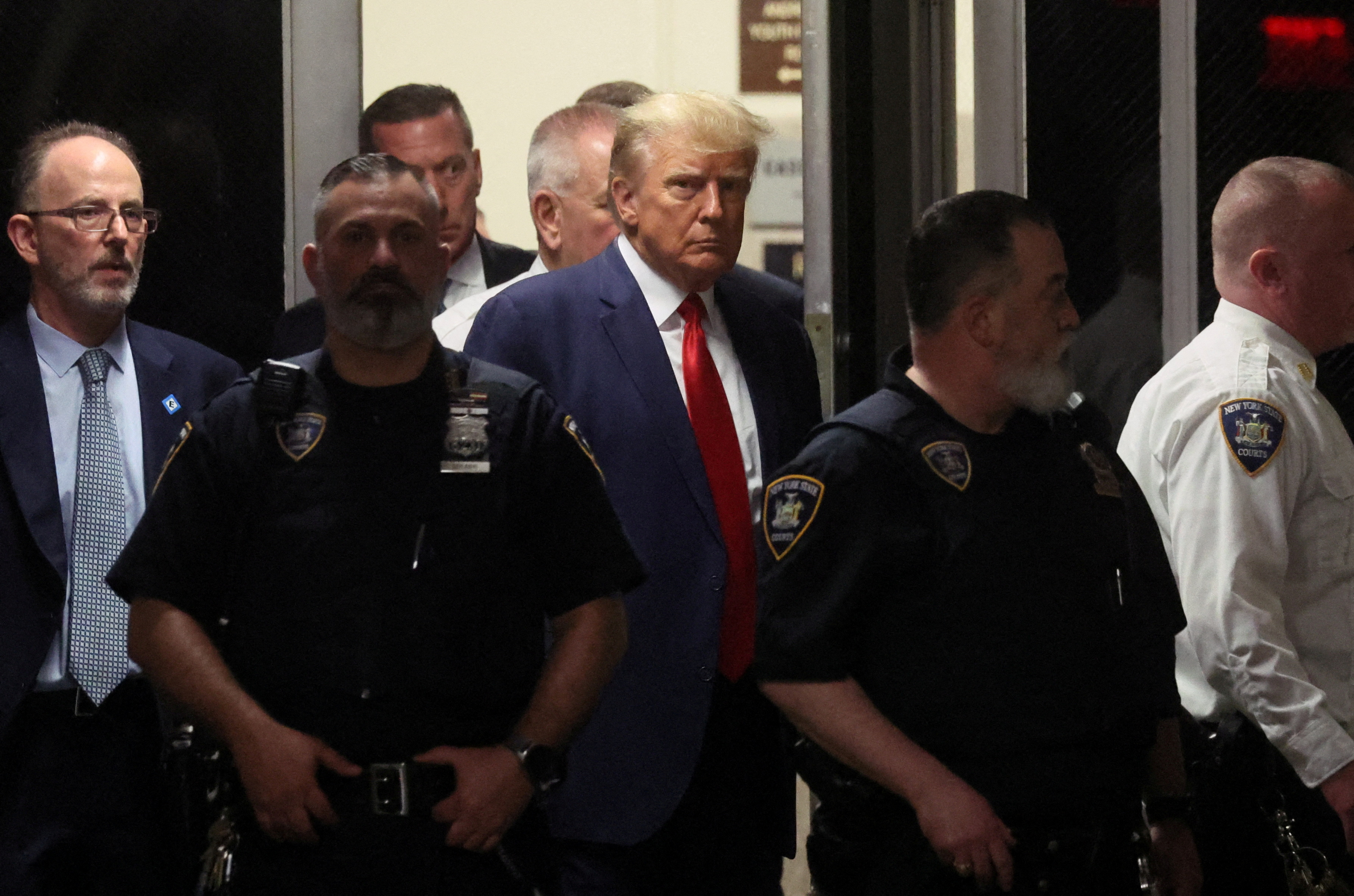 Former US President Donald Trump in a navy blue suit, being escorted by several police officers