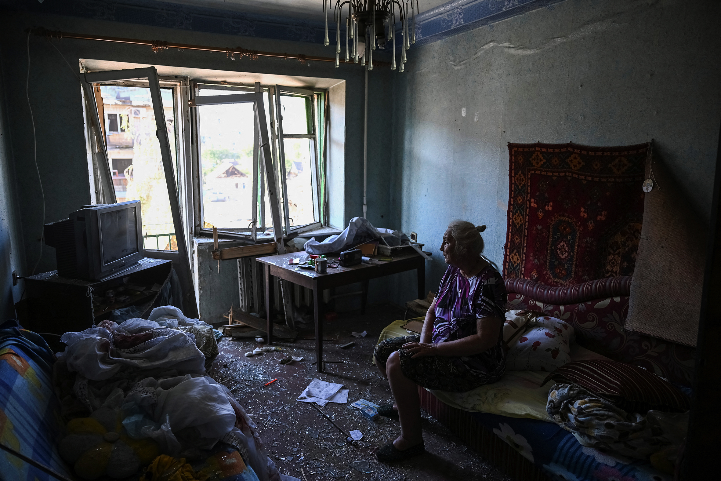 An elderly woman sits on a bed in her apartment. The windows have been smashed, and there is glass and rubble scattered across the floor.