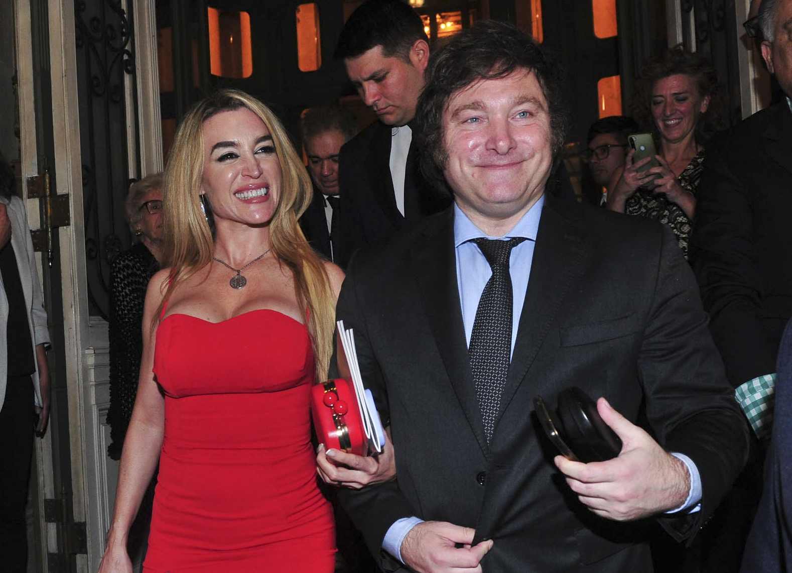 A man with sideburns and wearing a suit walks in arm with a woman in a red dress.