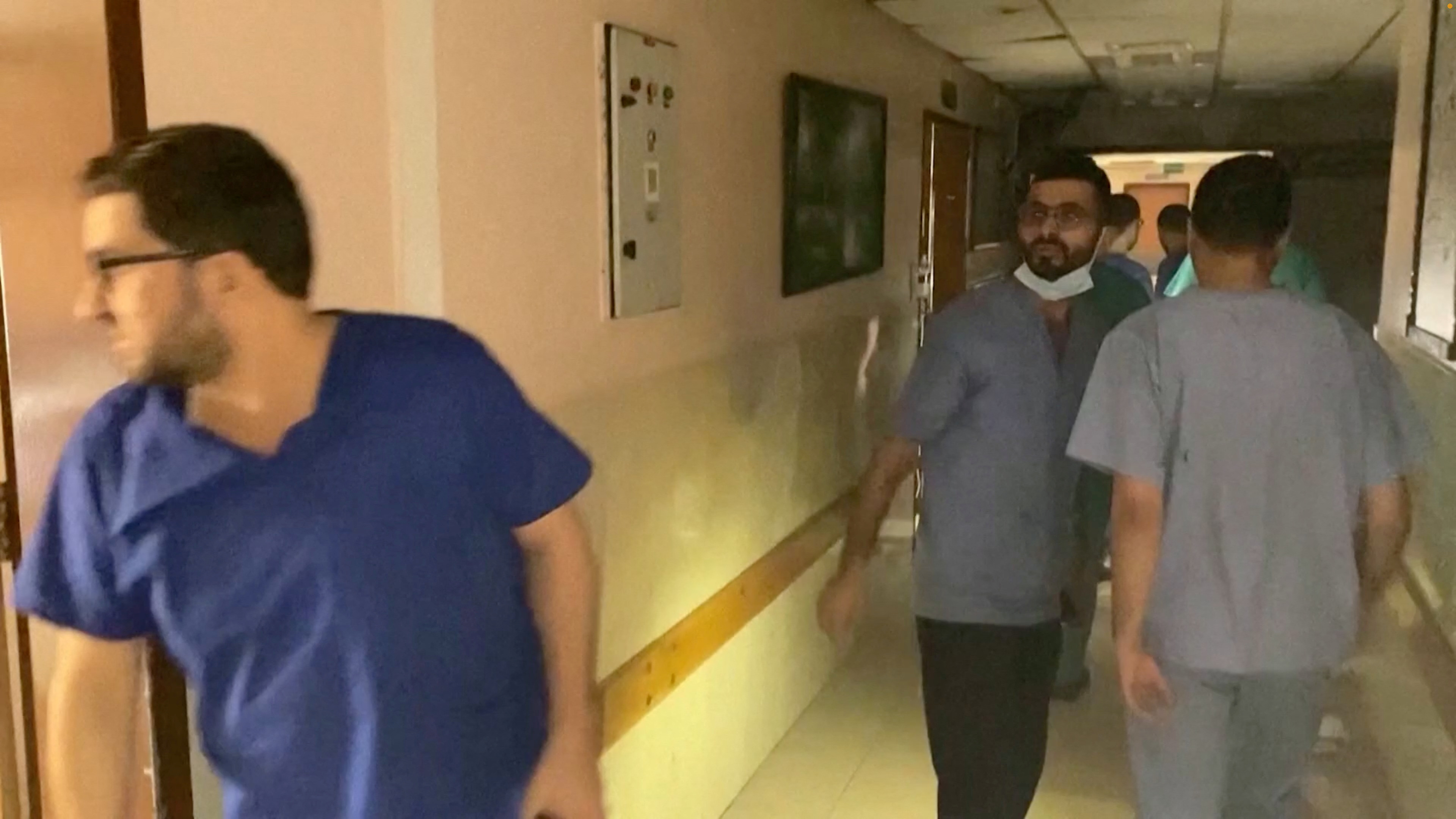 A doctor peers around a corridor, Other hospital staff stand in the darkened hallway behind him.