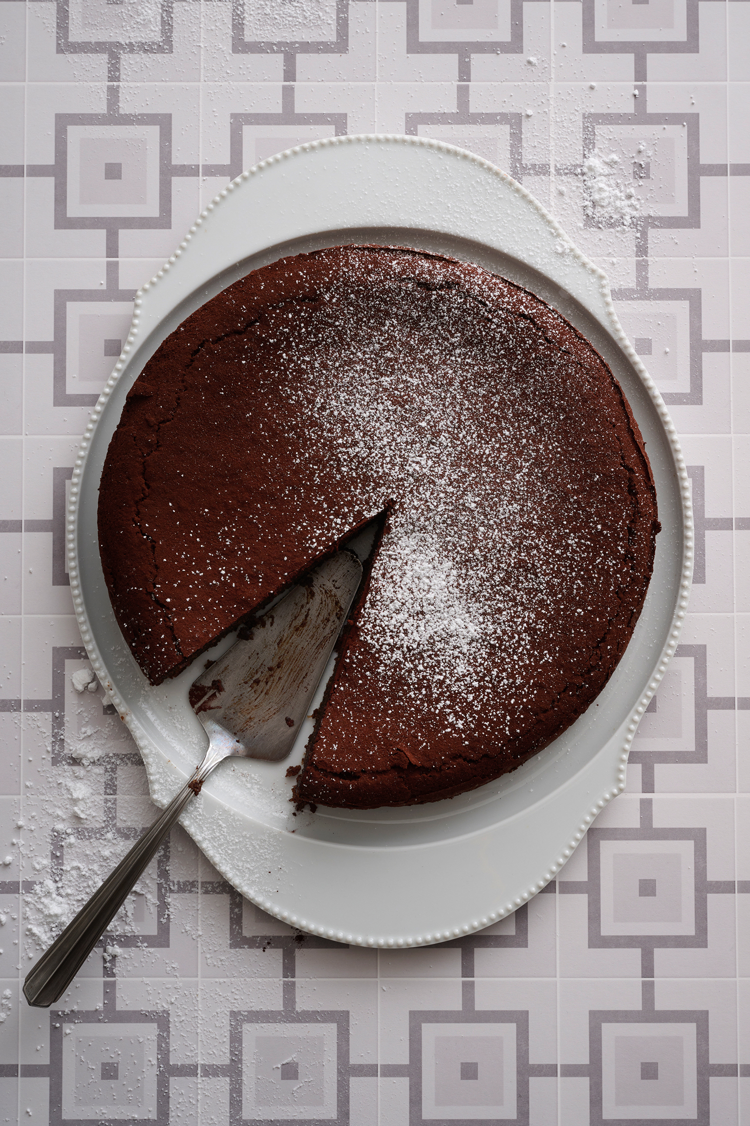 A chocolate cake sits on a plate with icing sugar and one slice removed from the centre
