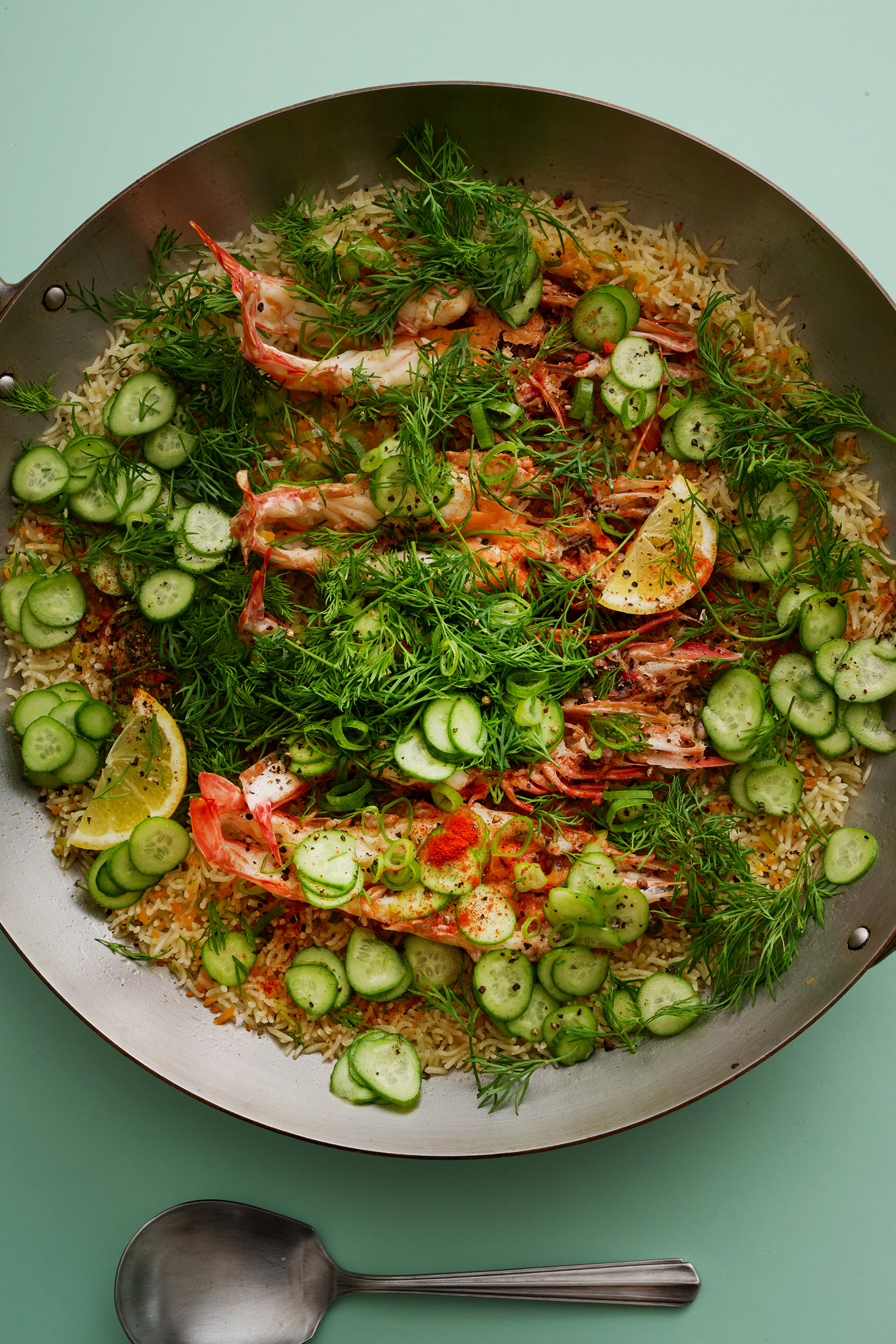 Rice, prawns, sliced lemon, cucumber, and a variety of herbs in a paella pan.