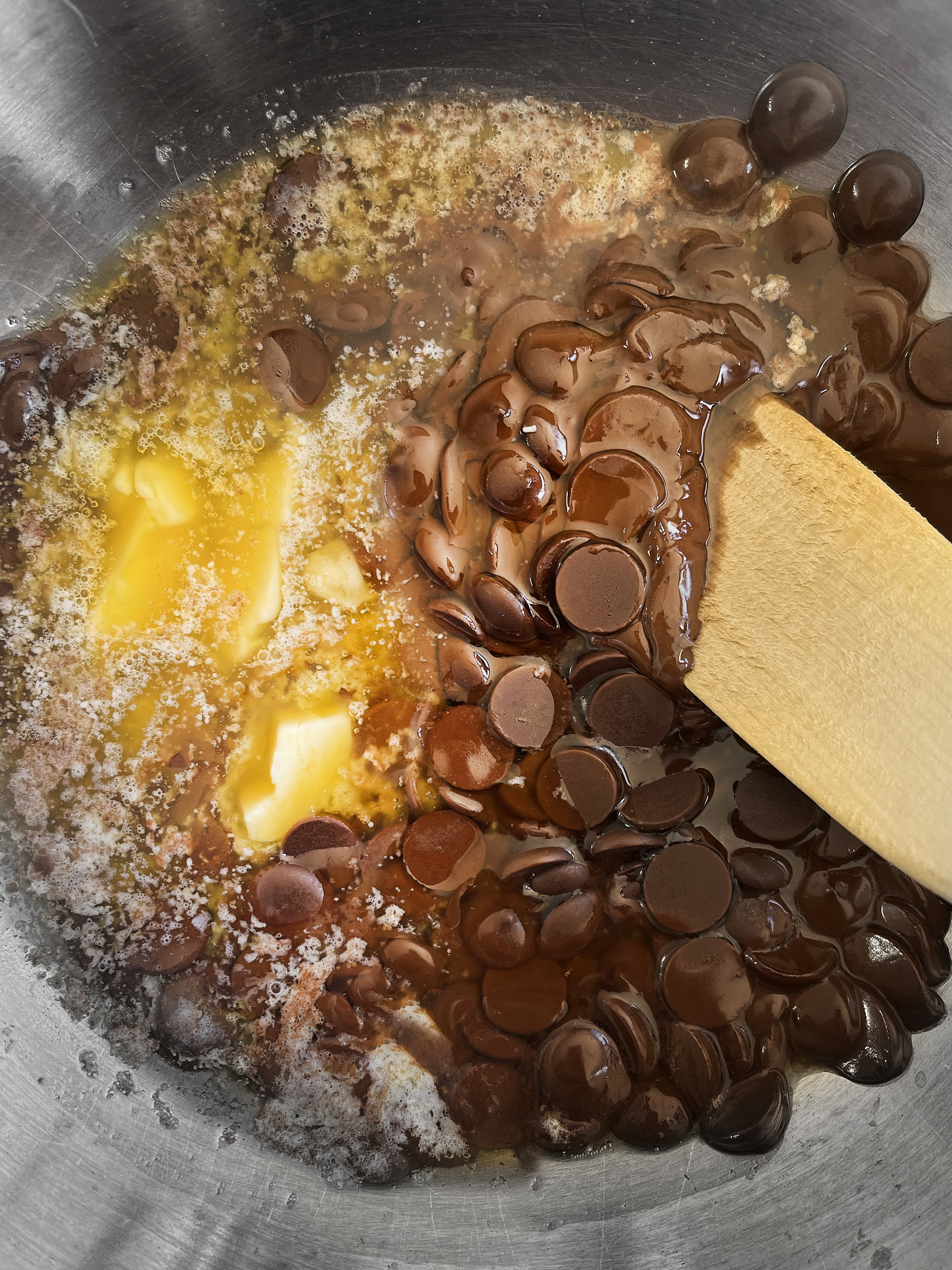 A silver mixing bowl holding chocolate chips, butter and sugar slowly melting into each other while being stirred by a wooden spatula