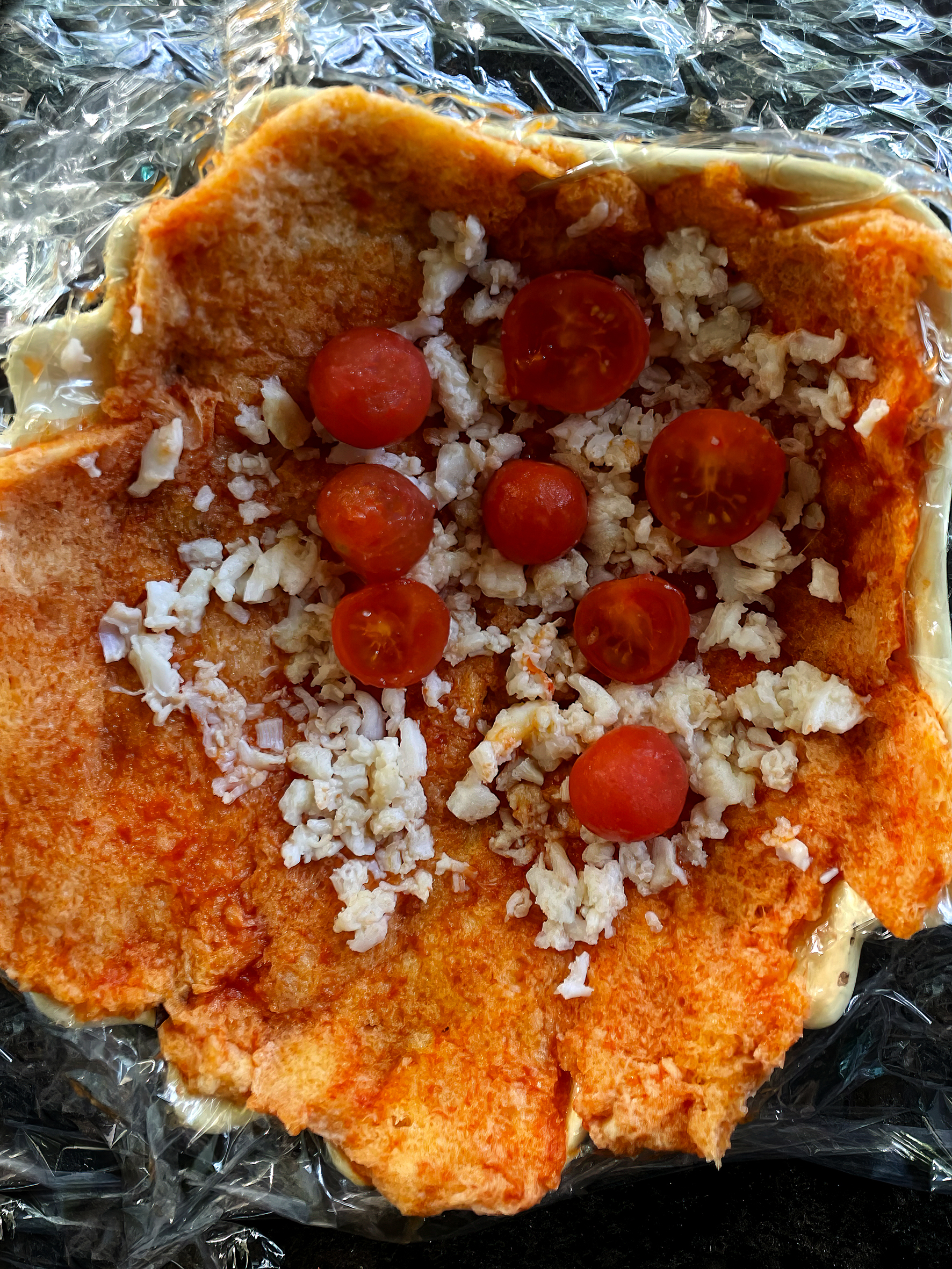 A circular bread shape holds chopped white onion and cherry tomato halves, surrounded by silver foil. It is in the prep stage and is not yet cooked.