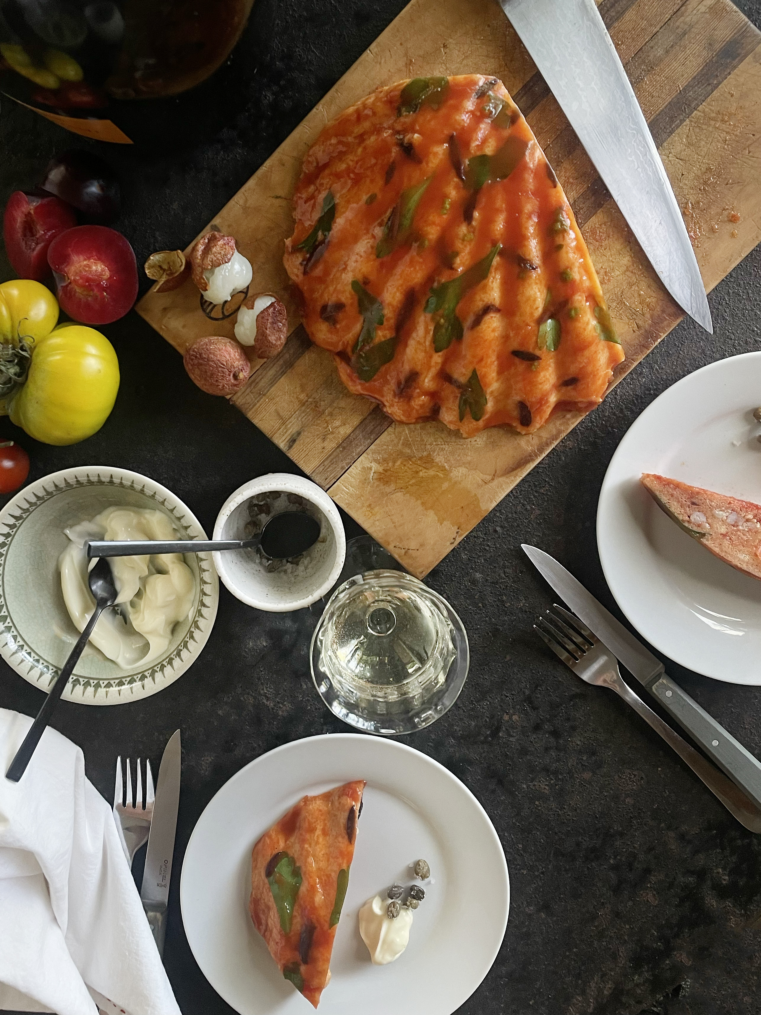 A kitchen bench holds a myriad of objects; a crab-shell shaped bread pudding sits at the top, sliced partially by a knife, with a portion on a white plate below, several glasses filled with wine and some yellow and red tomatoes off to the side