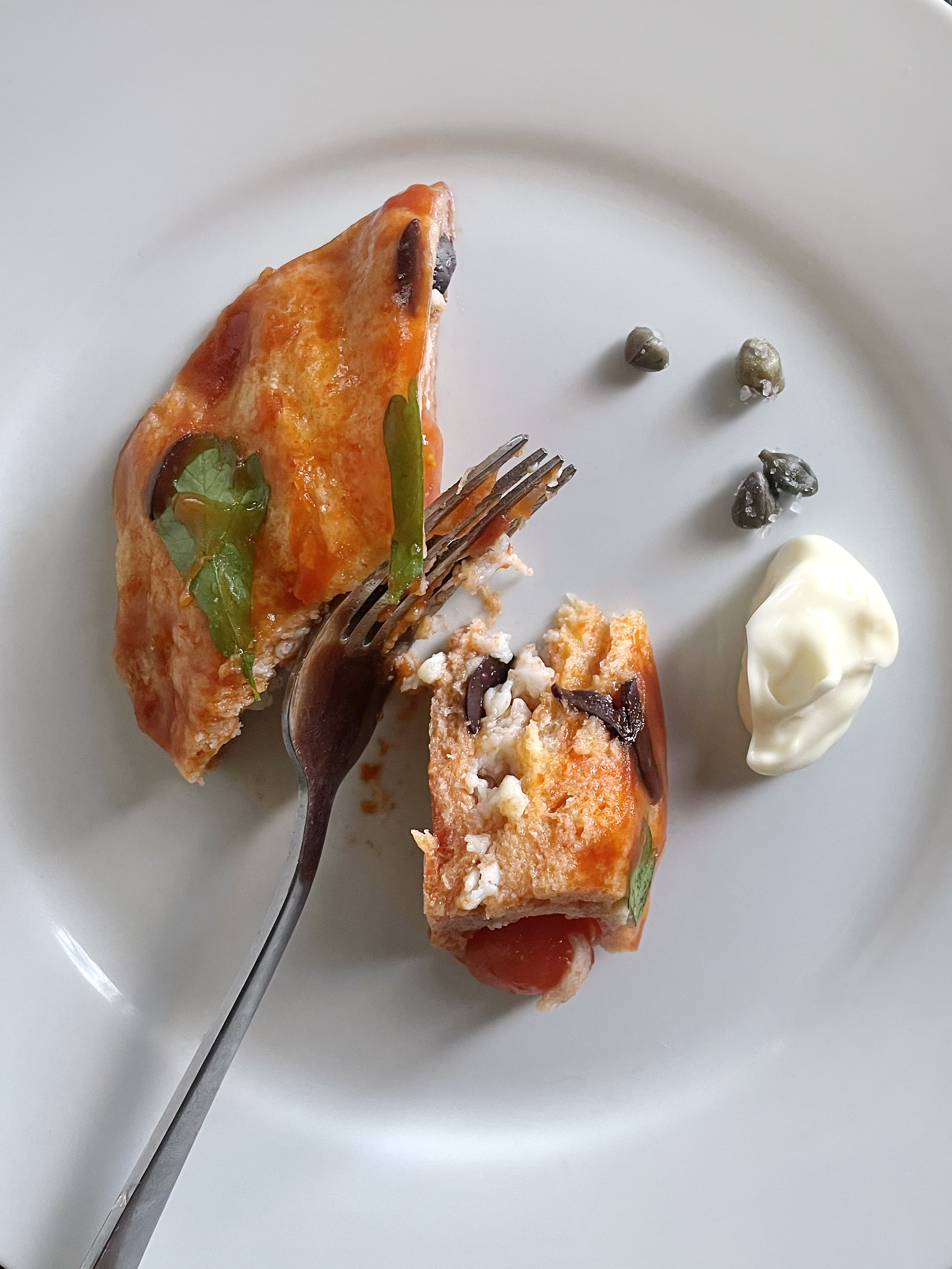 A white plate holds a singular slice of light brown-red crab pudding, with a basil leaf on top and a fork slicing off a small part