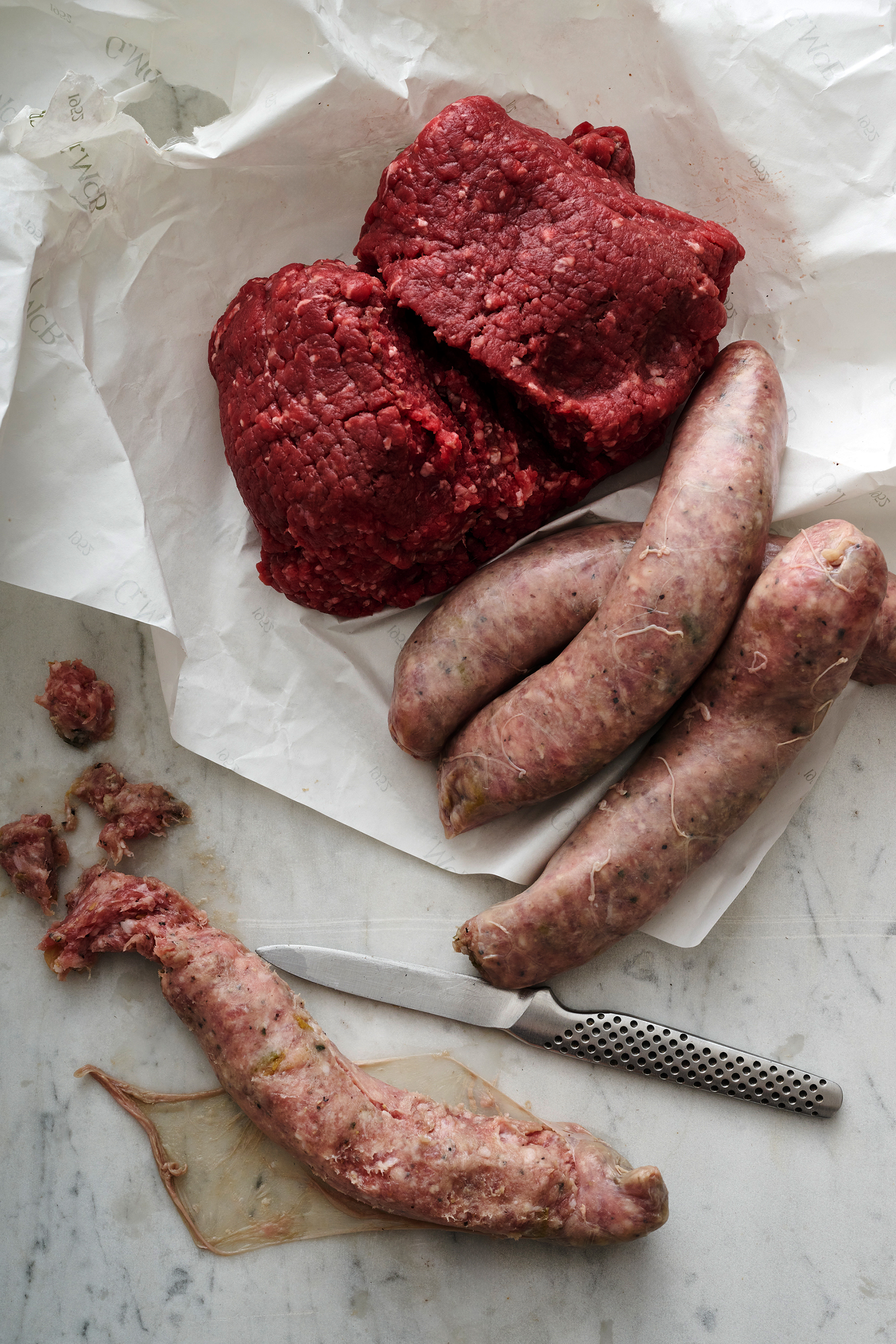 Raw beef mince and pork and fennel sausages sit on a piece of crinkled white butchers' paper, next to a paring knife.