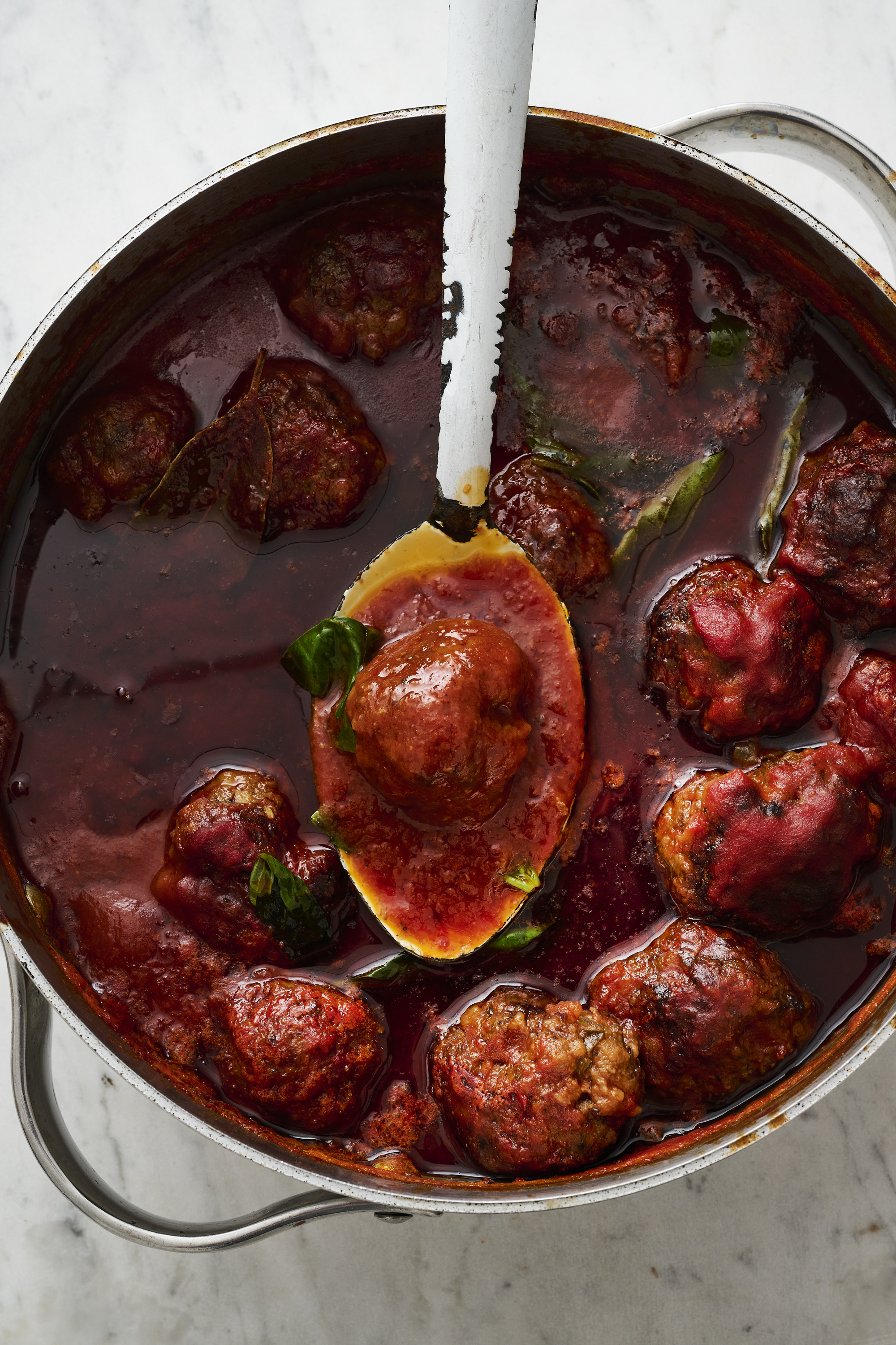 A big rustic pot of deep red meatballs in tomato sauce sits on a bench top, a silver spoon balancing on the pot's rim and holding a single meatball.