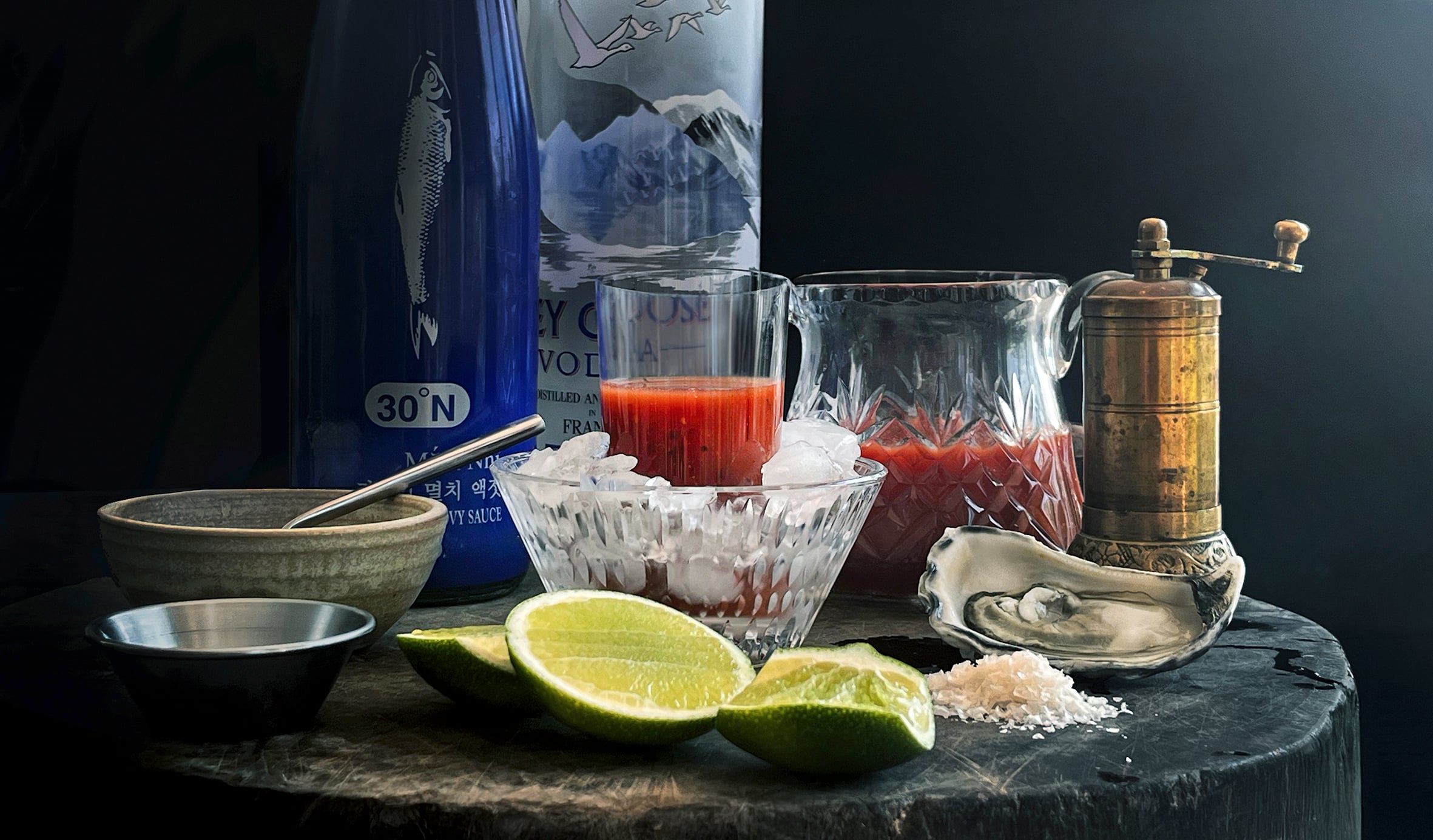 Ingredients for an oyster shot, including a halved lime, tomato juice, an oyster in its shell and a pepper grinder, sit on a table.