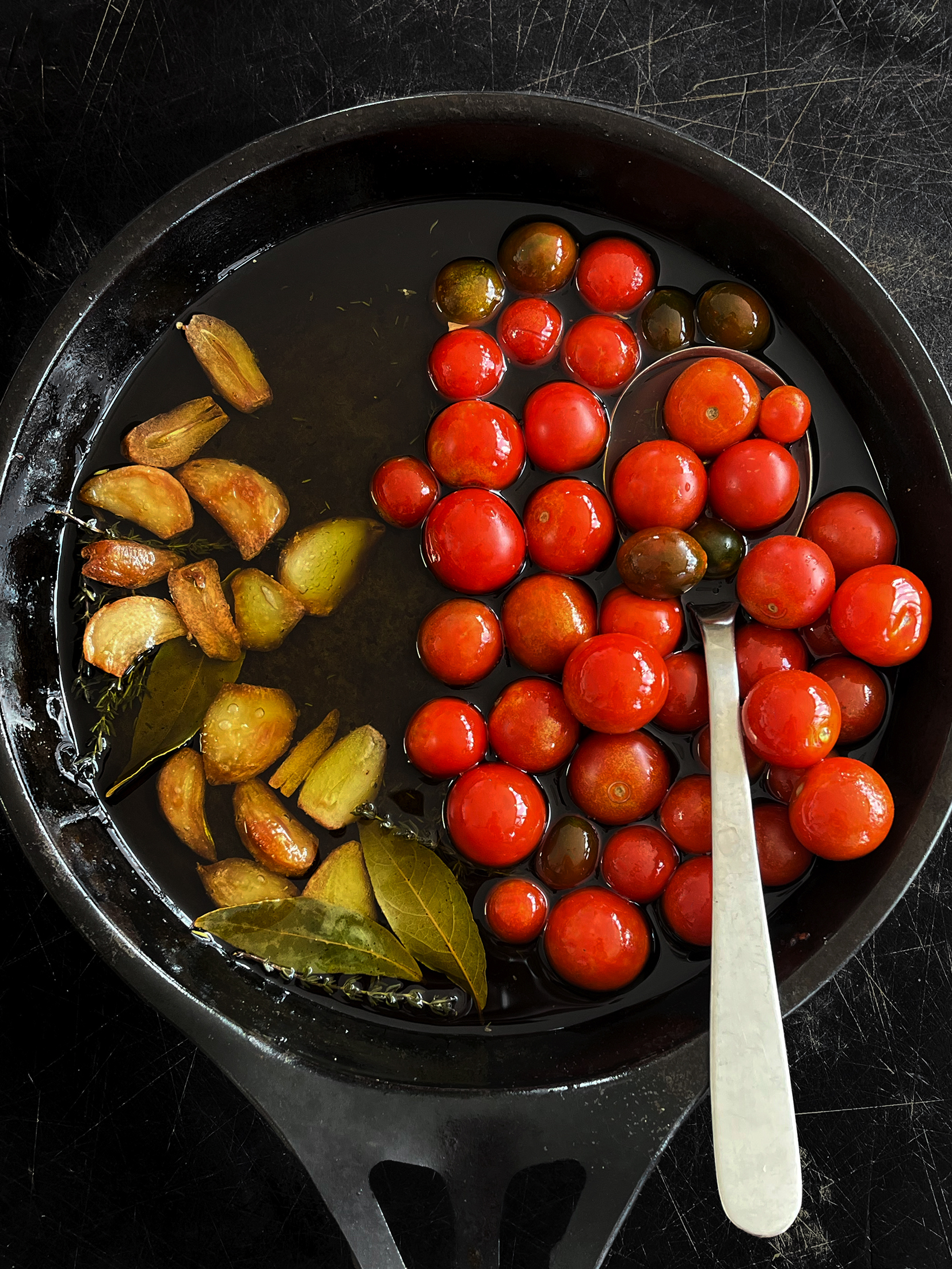 A wide-based saucepan filled with olive oil, bay leaves, thyme, kalamata olives, cherry tomatoes and cloves of garlic.